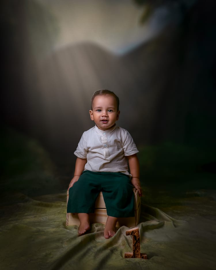 Niño pequeño sentado sobre una caja de madera, con camisa de lino blanca y pantalón verde oscuro, descalzo. A sus pies, un número uno tallado en madera. Fondo pictórico de bosque con haces de luz.