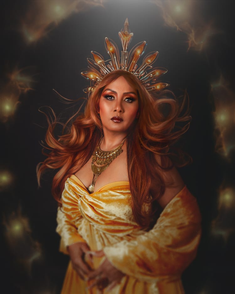 Retrato de estudio de una mujer con una corona radiante de cristal ámbar y el cabello cobrizo en movimiento. Viste terciopelo dorado y un collar de monedas. Luces cálidas difusas al fondo.
